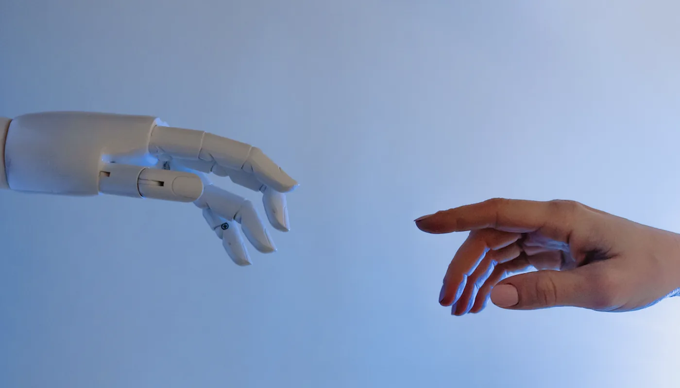 A white robotic hand and a human hand reaching toward each other against a pale blue background